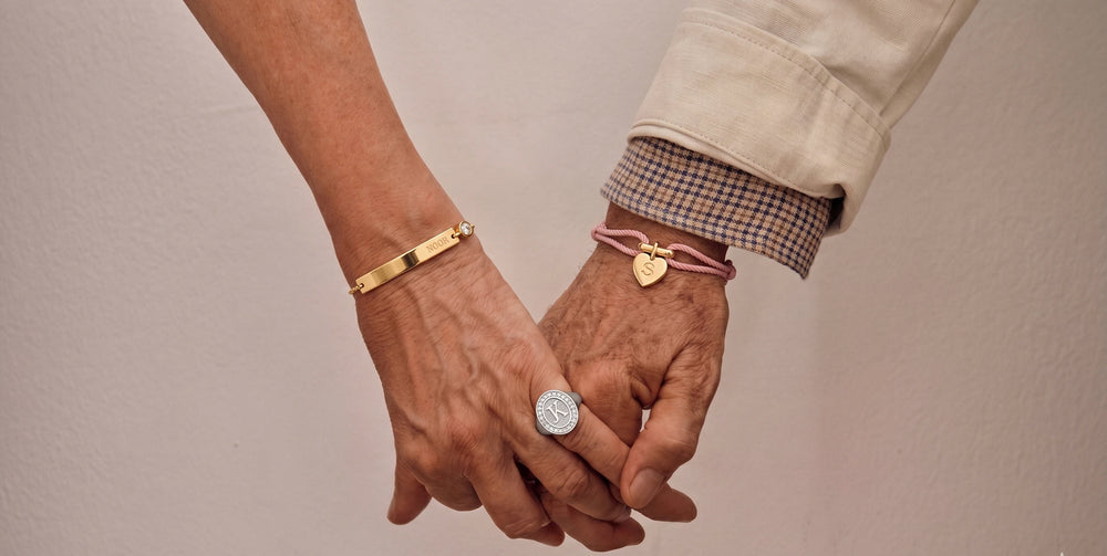 Two hands holding each other with visible jewelry on a neutral background
